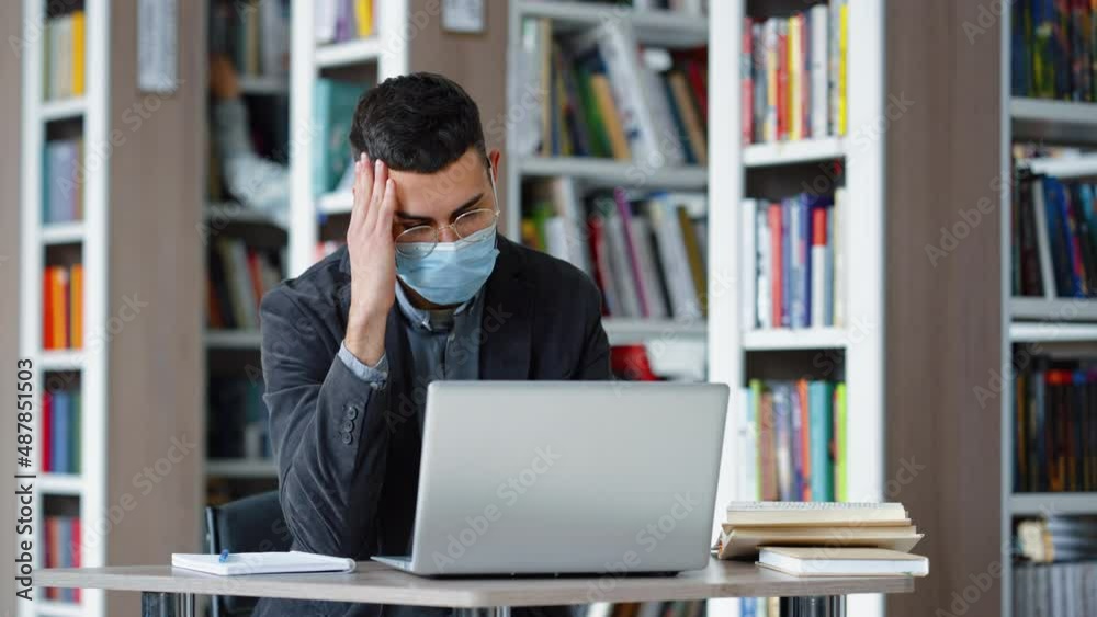 Student wearing eyeglasses and protective mask working on laptop in library, browsing internet, doing homework, blurred person on background taking book from bookshelf. Education at pandemic times Student wearing eyeglasses and protective mask working on laptop in library, browsing internet, doing homework, blurred person on background taking book from bookshelf. Education at pandemic times