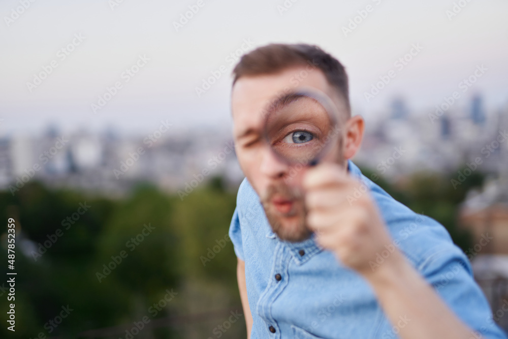 Attractive man closeup looking with magnifying glass at camera with amazed facial expression. Male wearing jeans shirt and peaked cap as detective or investigator concept. High quality image Attractive man closeup looking with magnifying glass at camera with amazed facial expression. Male wearing jeans shirt and peaked cap as detective or investigator concept. High quality image