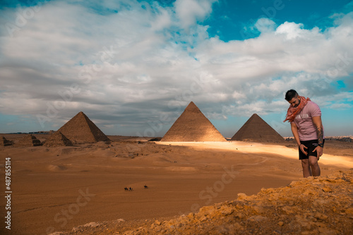 Man sitting on the sandy desert dunes posing in front of the great pyramids of giza. Traveling egypt in winter time, tourists posing for a picture