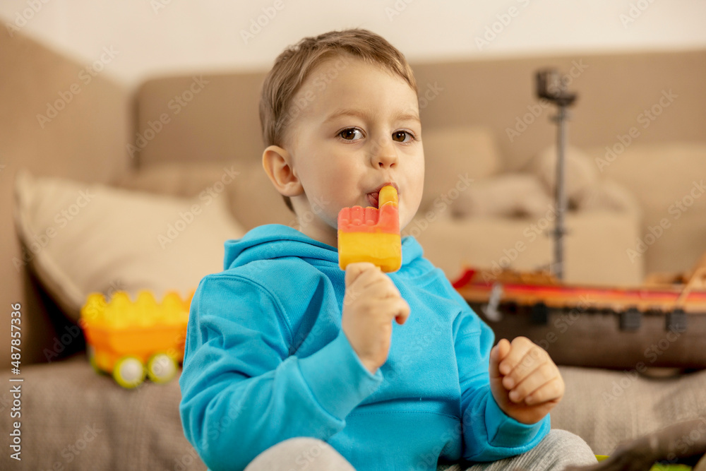 Little caucasian boy with blue hoodie playing with colourful toys and eating ice cream at home. Child having fun. Happy and cheerful kid plays with ship, dinosaurs, castle. Leisure activity. Little caucasian boy with blue hoodie playing with colourful toys and eating ice cream at home. Child having fun. Happy and cheerful kid plays with ship, dinosaurs, castle. Leisure activity.