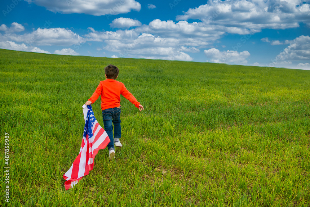 Child Running And Enjoying Himself In A Green Meadow With A Dramatic Sky. He Bears The Flag Of The United States, Wears A Red T-shirt And Jeans. Image With Copy Space. Nature And People. Child Running And Enjoying Himself In A Green Meadow With A Dramatic Sky. He Bears The Flag Of The United States, Wears A Red T-shirt And Jeans. Image With Copy Space. Nature And People.