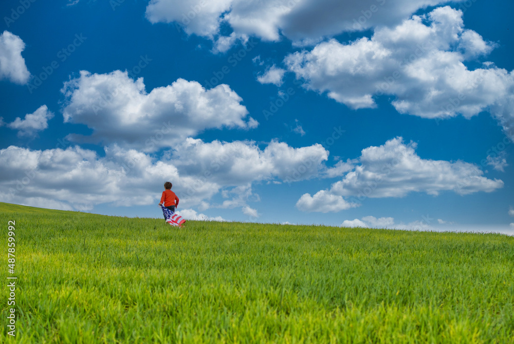 Child Running And Enjoying Himself In A Green Meadow With A Dramatic Sky. He Bears The Flag Of The United States, Wears A Red T-shirt And Jeans. Image With Copy Space. Nature And People. Child Running And Enjoying Himself In A Green Meadow With A Dramatic Sky. He Bears The Flag Of The United States, Wears A Red T-shirt And Jeans. Image With Copy Space. Nature And People.