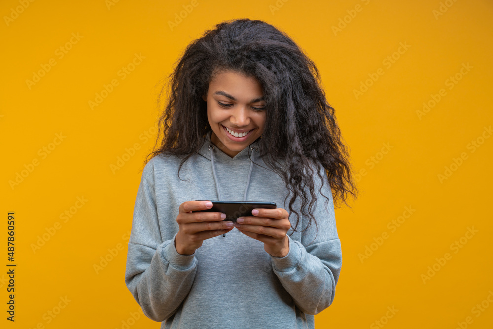 Studio portrait of beautiful casually dressed dark skinned girl with perfect charming smile texting message using her smartphone, isolated on bright colored yellow background Studio portrait of beautiful casually dressed dark skinned girl with perfect charming smile texting message using her smartphone, isolated on bright colored yellow background