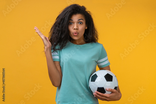 Portrait of young cute soccer fan girl holding a ball in hands, posing with excited face expression, isolated over bright colored yellow background © wpadington