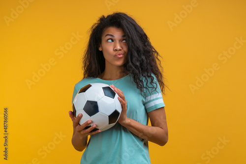 Studio portrait of cute cuquette soccer fan girl posing with a ball in hands, isolated over bright colored yellow background © wpadington