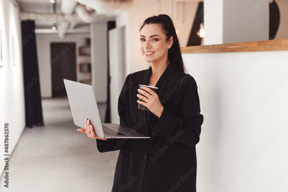 Young business woman with laptop and paper coffee cup standing in office Young business woman with laptop and paper coffee cup standing in office