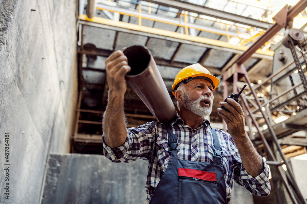 A senior factory worker relocates pipe and speaks with his coworker over the walkie-talkie. 