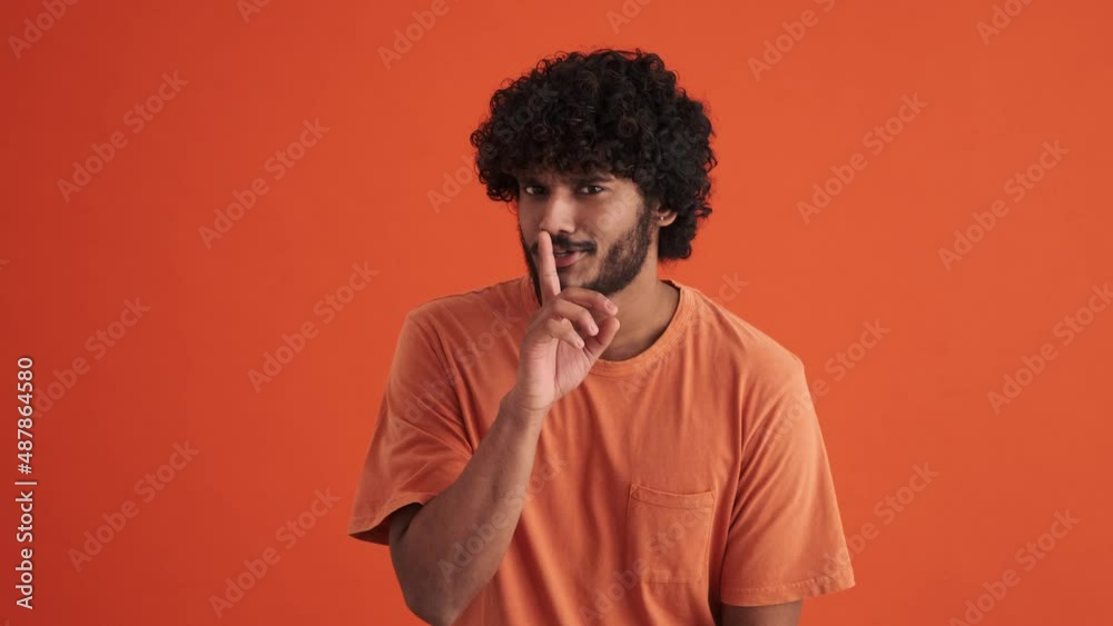 Positive curly-haired Indian man looking around and showing shh gesture in the orange studio Positive curly-haired Indian man looking around and showing shh gesture in the orange studio