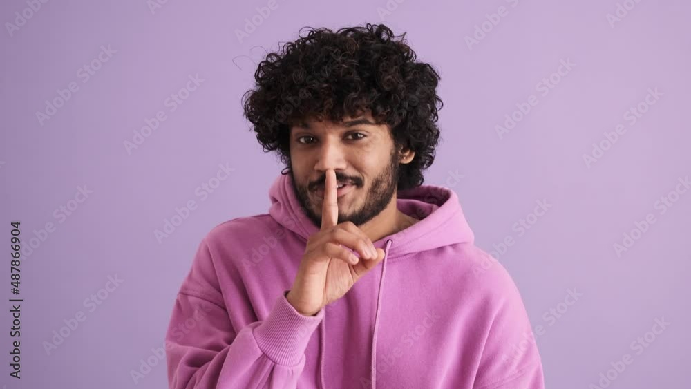 Happy curly-haired Indian man looking around and showing shh gesture in the purple studio Happy curly-haired Indian man looking around and showing shh gesture in the purple studio