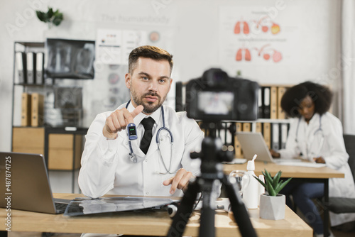 Caucasian medical specialist in white lab coat showing proper using of contactless thermometer while recording video on camera. Blog with usefell medical advices.