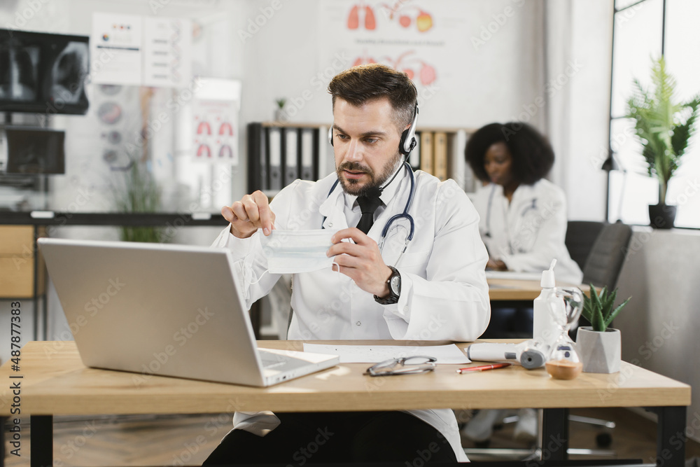 Caucasian male doctor with face mask in hands using headset and laptop for video call. African american nurse working at cabinet on background. Caucasian male doctor with face mask in hands using headset and laptop for video call. African american nurse working at cabinet on background.