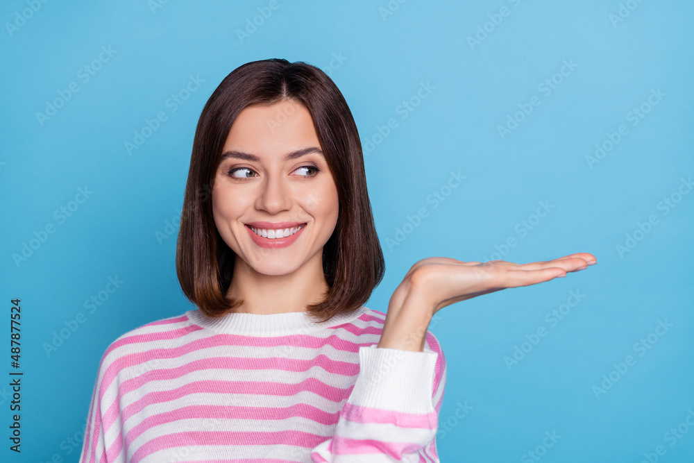 Photo of optimistic young lady look hold empty space wear striped shirt isolated on blue color background Photo of optimistic young lady look hold empty space wear striped shirt isolated on blue color background