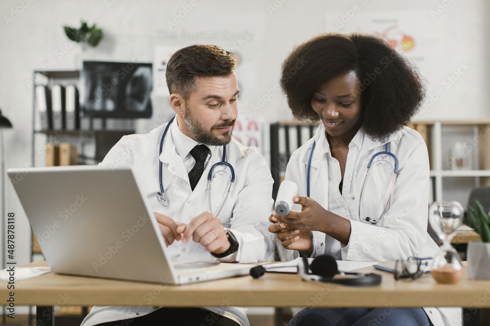 Multiracial male and female doctors using modern laptop for online seminar. Two medical workers sitting at desk and talking about usage of contactless infrared thermometer. Multiracial male and female doctors using modern laptop for online seminar. Two medical workers sitting at desk and talking about usage of contactless infrared thermometer.