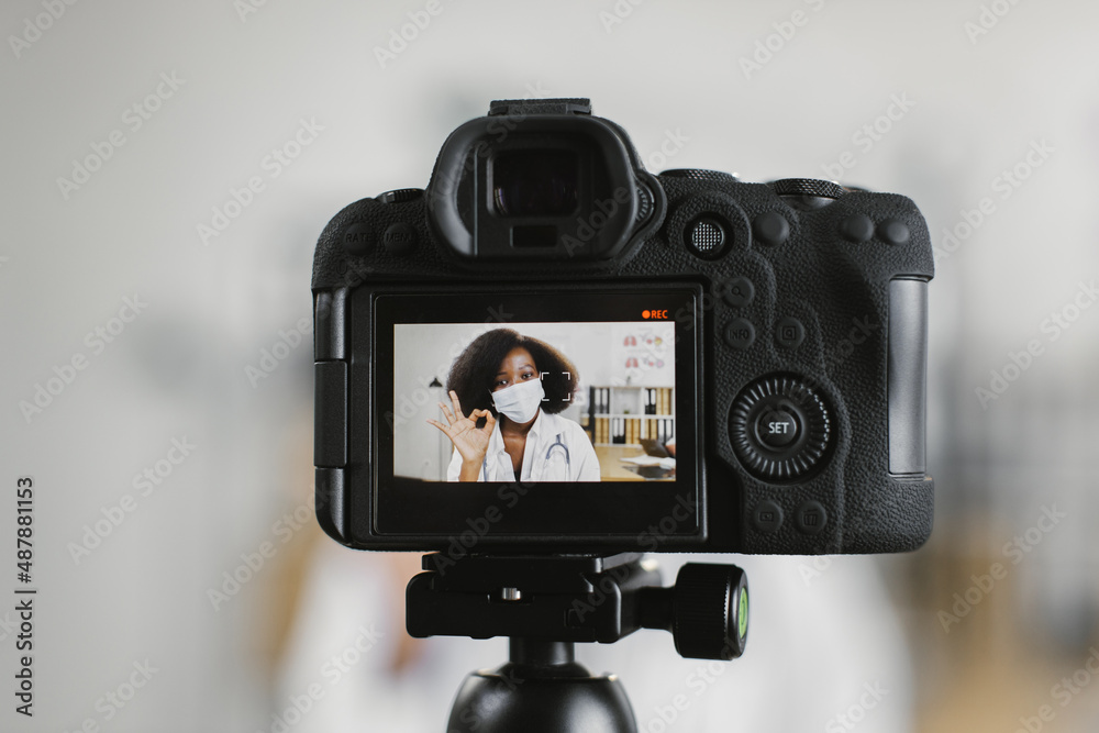 Video camera screen with confident african american female doctor in white lab coat and face mask. Competent medical worker talking and gesturing during video recording.