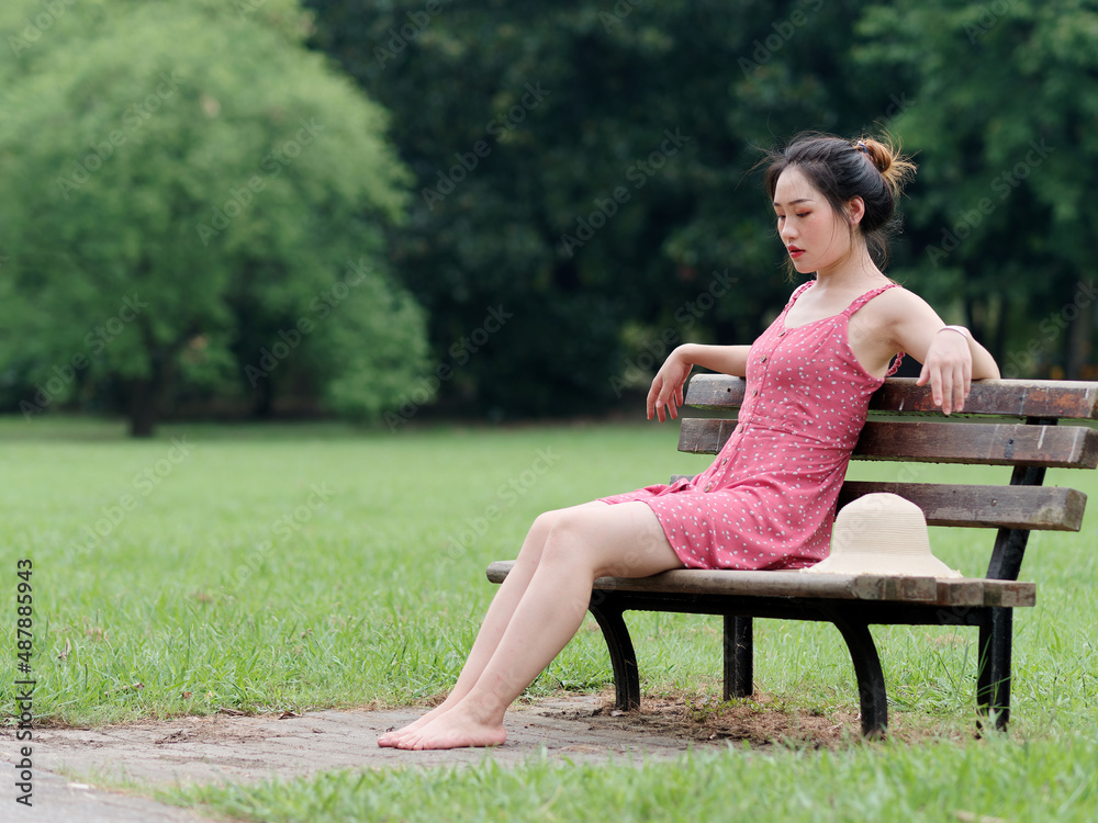 Portrait of young Asian woman sitting on bench on grass field with barefoot in forest park, beautiful Chinese girl in sexy red dress enjoy her carefree time in hot summer day, full length shot.