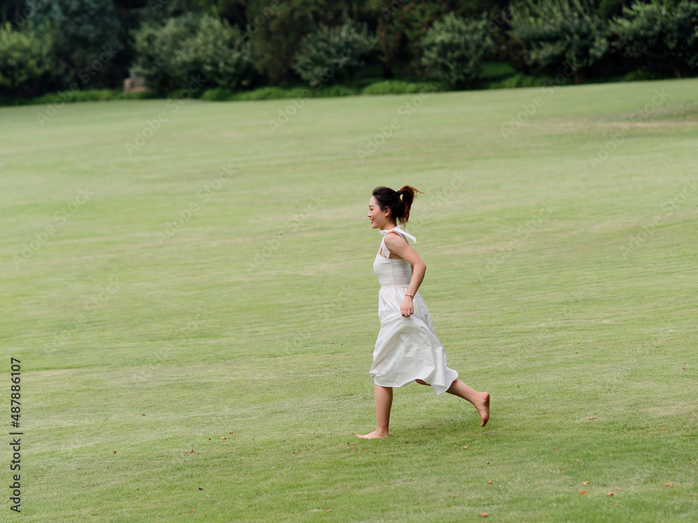 Portrait of young Asian woman running on grass field with barefoot in forest park, beautiful Chinese girl in white dress enjoy her carefree time in sunny summer day, full length shot.