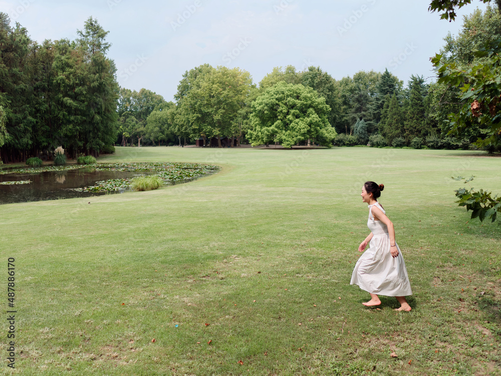 Portrait of young Asian woman running on grass field with barefoot in forest park, beautiful Chinese girl in white dress enjoy her carefree time in sunny summer day, full length shot.