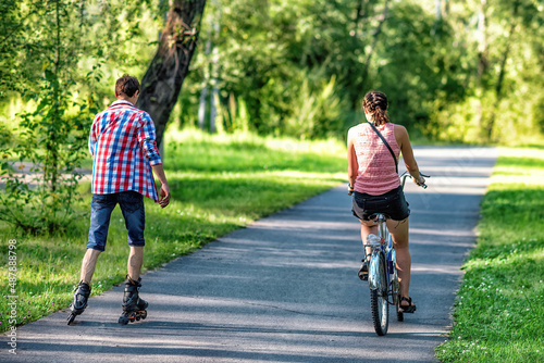 Two friends riding a bicycle and roller skating in a park in summer.