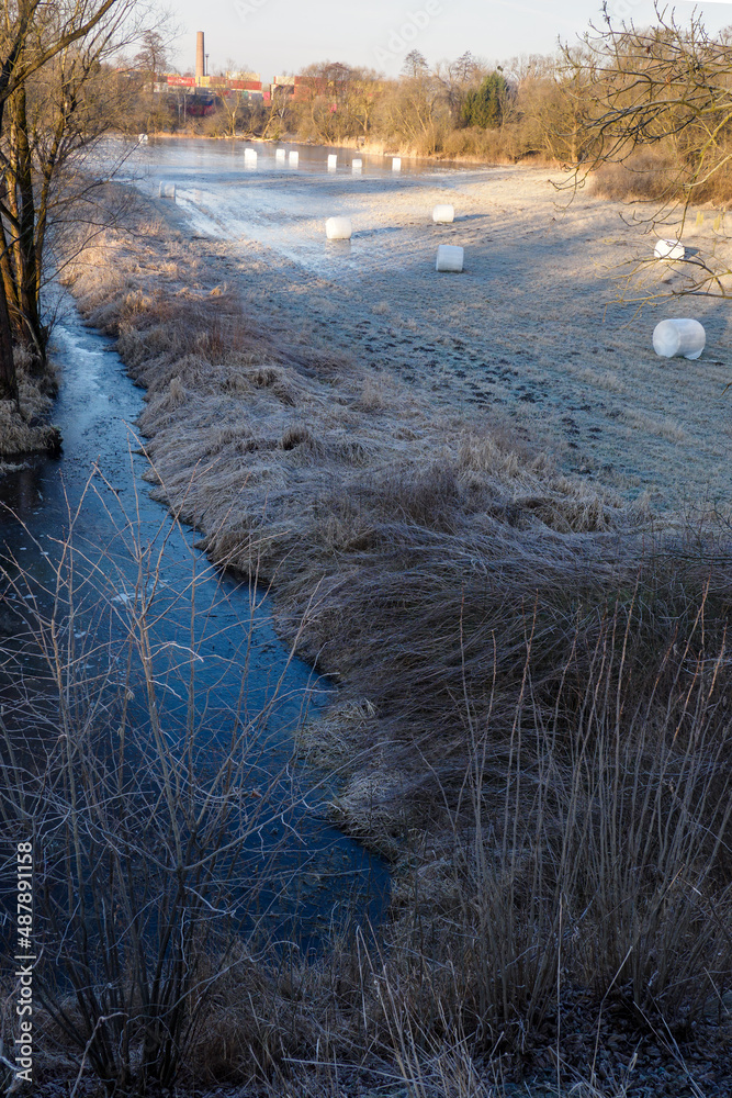 A flowing river with a frozen flooded meadow and white bales.