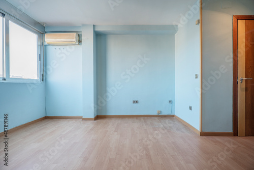 Image of a studio apartment with blue painted walls and a wooden floor and a window with an air conditioner on the wall