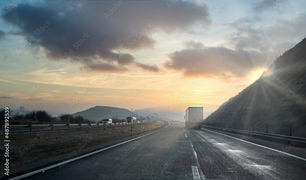 a wet freeway at dawn on a cloudy day, with mountains in the background and trucks driving in both directions, sunbeams behind the mountains