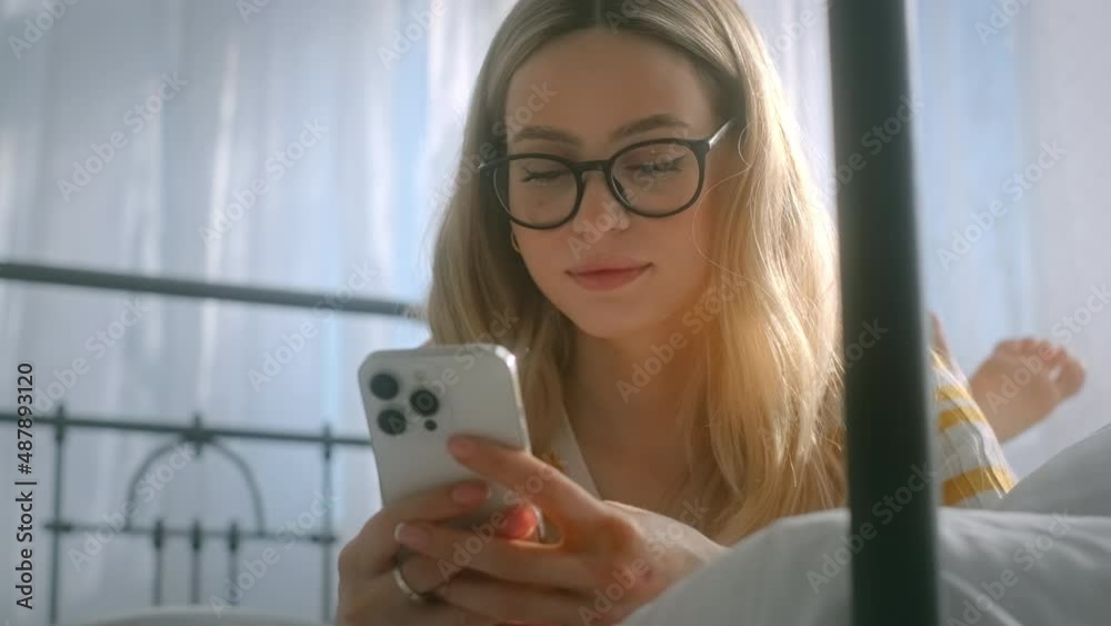 Happy smiling young attractive caucasian woman in eyeglasses using smartphone, lying on a bed in sunny day. Happy smiling young attractive caucasian woman in eyeglasses using smartphone, lying on a bed in sunny day.