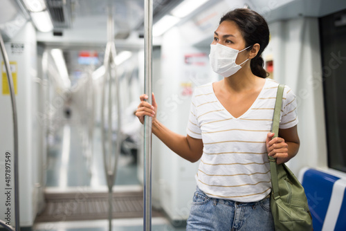 Asian woman in face mask with shoulder bag standing in subway train and holding handrail.
