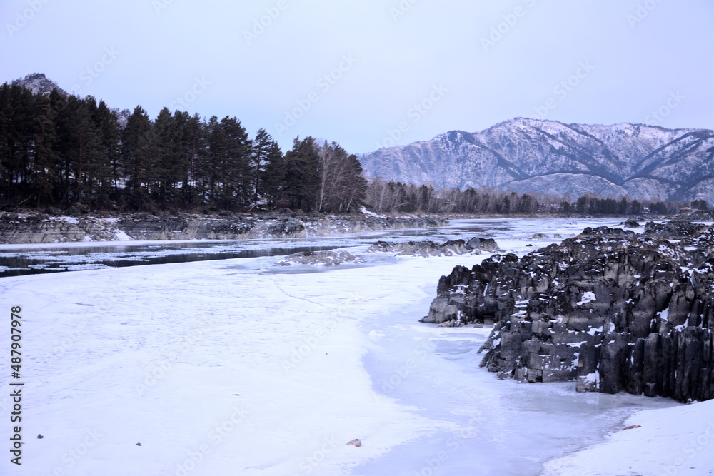 Rocky banks of a frozen river, a strip of pine forest and snow-capped mountains in the early morning.