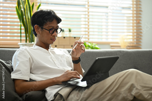 Asian man in casual clothes sitting on couch and working online with computer tablet.