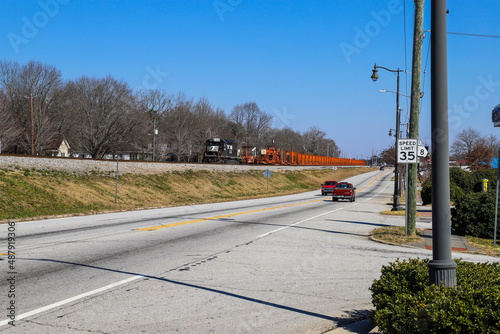 a shot of a black and orange train on the tracks near a street with cars driving with yellow and green grass and brown and bare winter trees along the track with blue sky in Douglasville Georgia USA photo