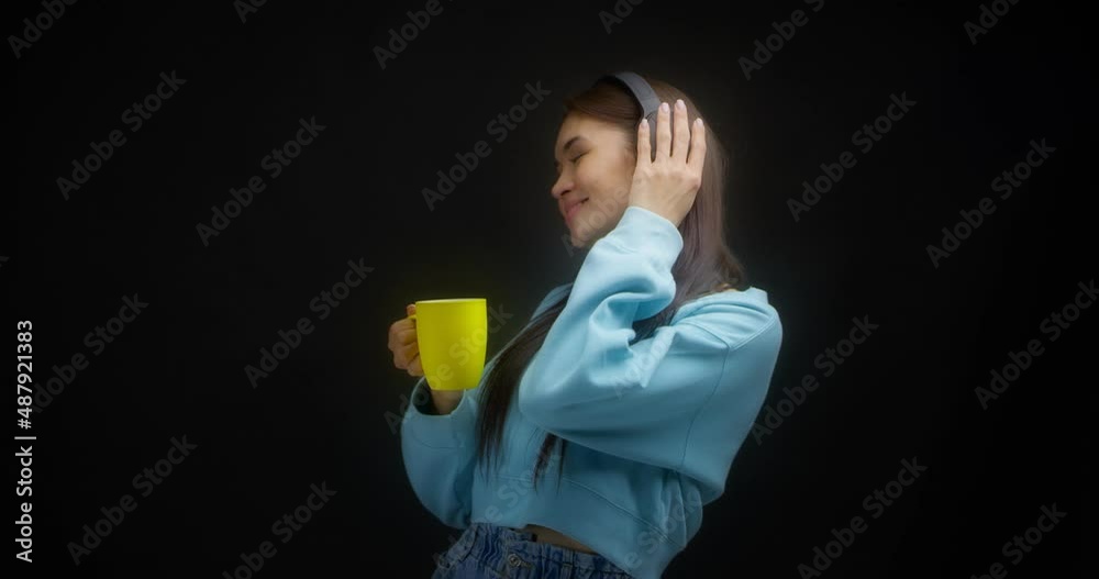 Cute young woman in a bright blue sweatshirt enjoys music in wireless headphones and drinks from a yellow mug. Asian woman dancing to the song isolated on a black background. Cute young woman in a bright blue sweatshirt enjoys music in wireless headphones and drinks from a yellow mug. Asian woman dancing to the song isolated on a black background.