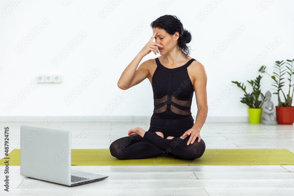 Fototapeta premium Online yoga lesson. Young yogi woman practices breathing yoga in yoga studio with laptop.
