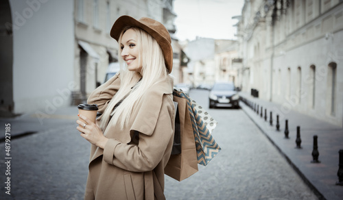 Young attractive blonde woman in autumn coat and hat holding lot of shopping packages and coffee cup in European street. Travel and shopping concept