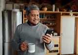 Happy multi-cultural elderly male typing a message on smartphone while standing in modern kitchen holding morning coffee.
