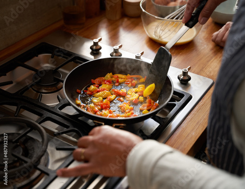 Multi-cultural elderly female frying vegetables with spatula in pan at modern stove.