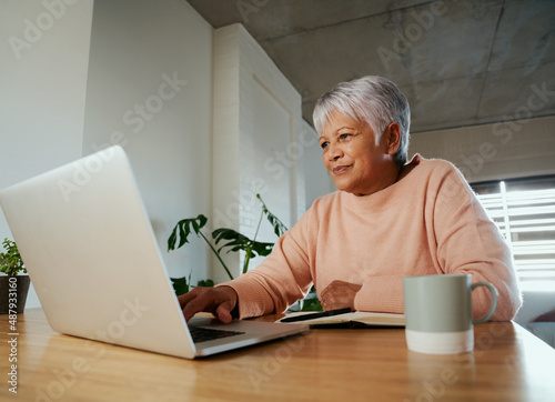 Elderly multi-ethnic female sitting at home counter scrolling on her laptop, smiling.