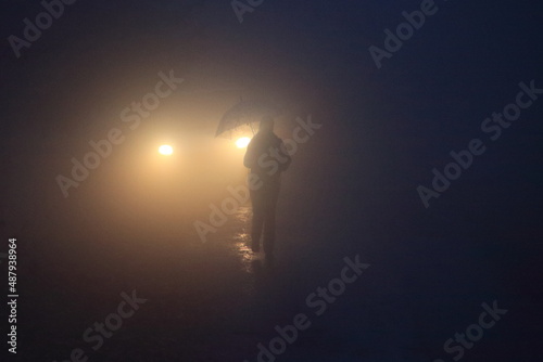 Foggy person with umbrella in front of car lights