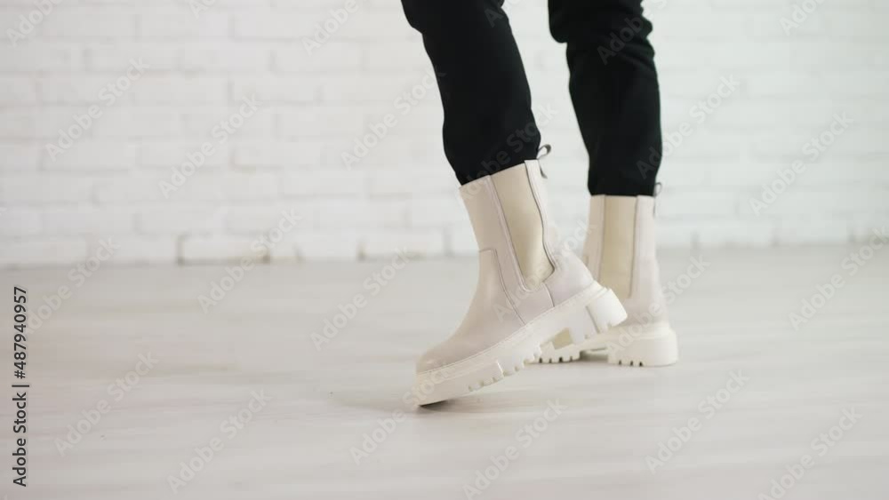 Woman in black jeans walks in new stylish white boots. Demonstrating the shoes of top fashion. White brick wall backdrop. Woman in black jeans walks in new stylish white boots. Demonstrating the shoes of top fashion. White brick wall backdrop.