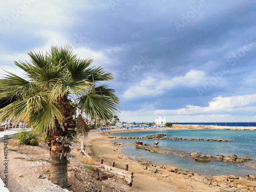 A palm tree, a sandy shore with stones, a beach with sun loungers and parasols, the Church of St. Nicholas the Wonderworker against a dramatic sky. © Elena