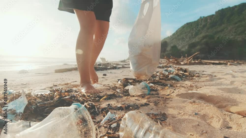 Eco-activist and volunteer walks barefoot on sea sand and collects discarded plastic bottles in bag for further disposal or reuse, showing environmental consciousness and worrying about climate change
