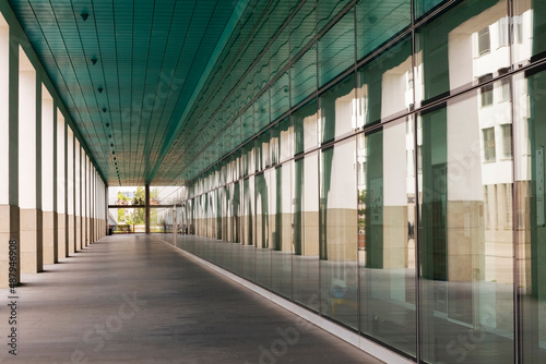 Architecture of a building with an outdoors hallway, leading the eye down this hallway to the end where vegetation can be seen. Reflections in the large windows also add mood to the image.
