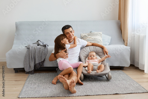 Happy father playing with his daughters, little girl hugging dad, man wearing white casual style T-shirt and jeans short sitting on floor with newborn baby and elder kid.
