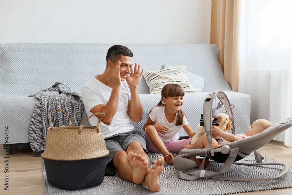 Portrait of funny man wearing white casual style T-shirt and jeans short sitting on floor with his children, father playing peek-a-boo with newborn baby in rocking chair.
