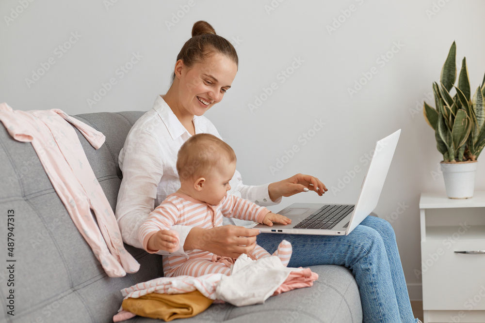 Indoor shot of dark haired woman freelancer wearing white shirt and jeans sitting on sofa with baby daughter and working on notebook, looking smiling at child, maternity leave and online job.