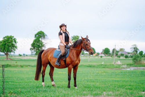 Glorious sport cowgirl horse woman with hat, holding harness, training. Horseback riding dressage, sky, meadow farm ranch outside. Brown black white horse, blue saddle. Oneness with nature, grass