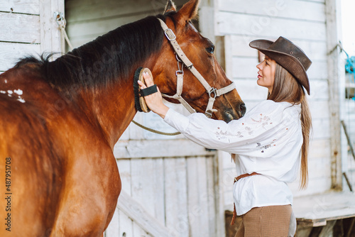 Fototapeta Naklejka Na Ścianę i Meble -  Tender horsewoman equestrienne in hat, trousers, shirt, belt. Cleaning, grooming horse mane. Animal care. Horse back riding. Bound between owner and horse. Outdoor farm, stall, stable. Side view
