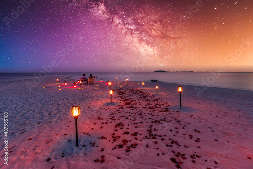 Amazing beach dinner setting under Milky Ways night sky. Luxury destination dining, honeymoon or anniversary dinner, flowers and candles for the best romantic experience. Stunning colorful outdoors photo