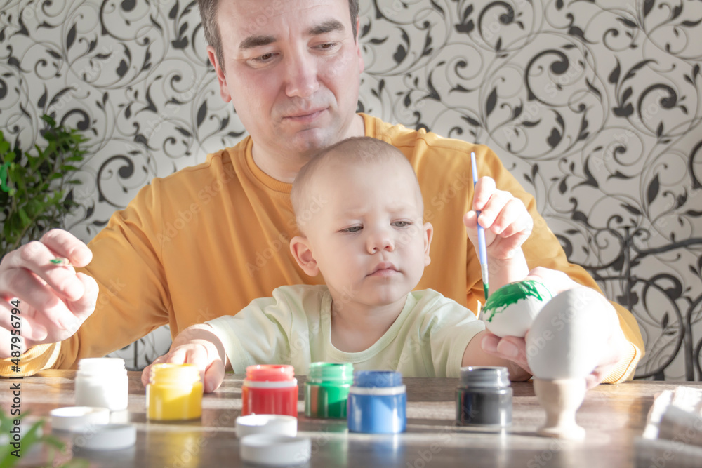 A dad and a little boy prepare for Easter at home indoors, paint Easter eggs with early-colored paints, sit at a table, sunlight falls on a wooden table. A dad and a little boy prepare for Easter at home indoors, paint Easter eggs with early-colored paints, sit at a table, sunlight falls on a wooden table.