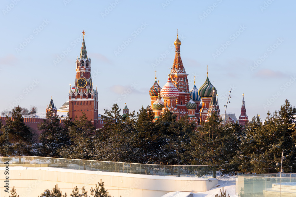 Fototapeta premium A winter shot of the Moscow Kremlin and St. Basil's Cathedral in the background and the coniferous trees of Zaryadye Park in the foreground. Snow on the ground. Famous tourist places