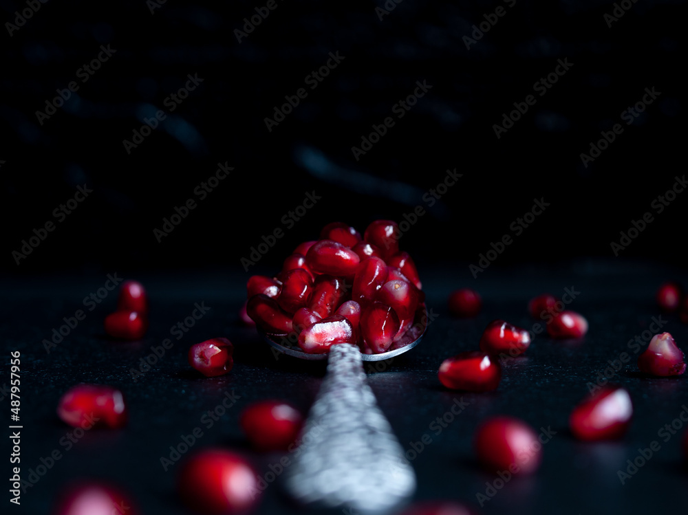Red pomegranate in breakfast in dark key, on black background with a silver spoon Red pomegranate in breakfast in dark key, on black background with a silver spoon